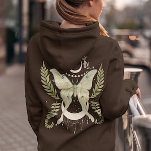 Puede incluir: Una sudadera marr&oacute;n con un estampado gr&aacute;fico de una polilla blanca con hojas verdes y una luna creciente en la espalda.