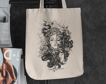 May include: A beige tote bag with a black and white illustration of a woman's face with a crown of thorns and the text "Embrace Your Darkness".