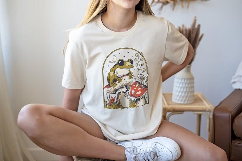 May include: A cream-colored t-shirt with a graphic of a green frog sitting on a mushroom. The mushroom has red and white accents. The graphic is surrounded by a crescent moon, stars, and a green vine.