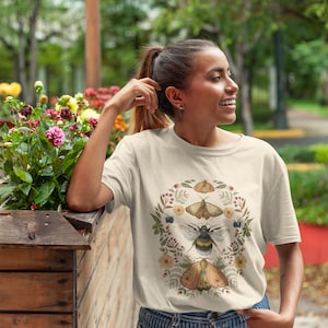 K&ouml;nnte beinhalten: Ein cremefarbenes T-Shirt mit einem floralen Kranz-Design mit einer Biene, einem Schmetterling und Motten. Der Kranz besteht aus kleinen Blumen, Beeren und Bl&auml;ttern.