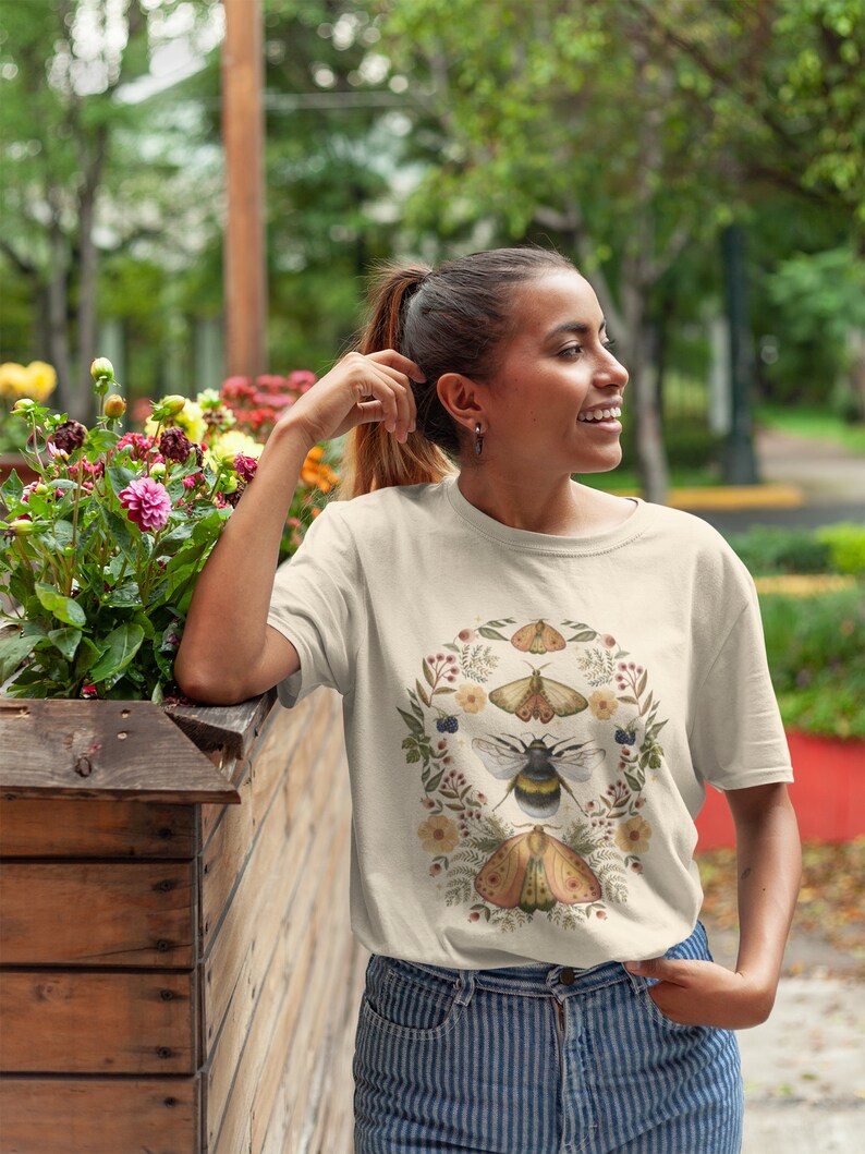 K&ouml;nnte beinhalten: Ein cremefarbenes T-Shirt mit einem floralen Kranz-Design mit einer Biene, einem Schmetterling und Motten. Der Kranz besteht aus kleinen Blumen, Beeren und Bl&auml;ttern.
