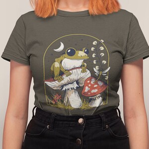 May include: A dark green t-shirt with a graphic of a green frog sitting on a mushroom with a red mushroom in the background. The graphic is surrounded by a gold frame with a crescent moon and stars.