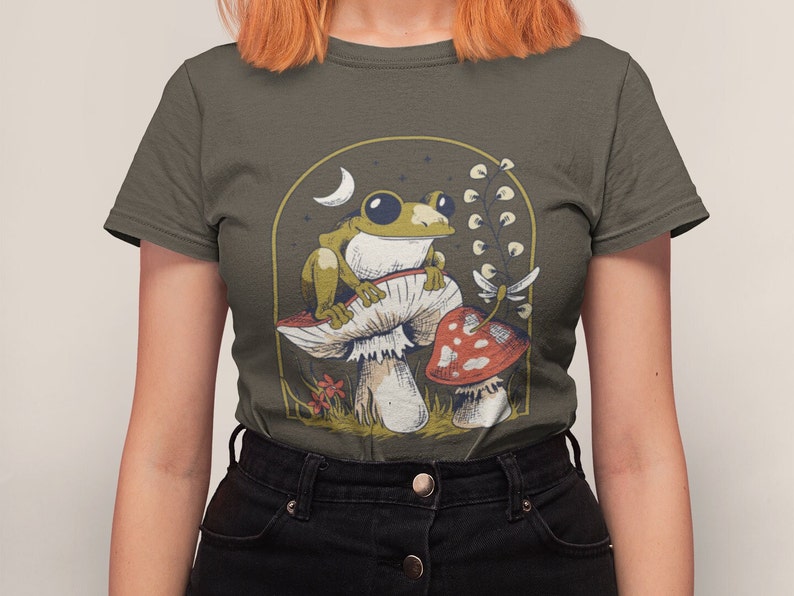 May include: A dark green t-shirt with a graphic of a green frog sitting on a mushroom with a red mushroom in the background. The graphic is surrounded by a gold frame with a crescent moon and stars.