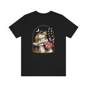 May include: Black t-shirt with a graphic of a green frog sitting on a mushroom with a red mushroom in the background.
