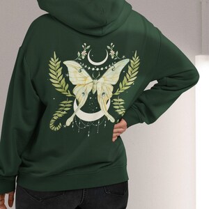 Puede incluir: Una sudadera con capucha verde oscuro con un dise&ntilde;o de polilla luna blanca en la espalda. La polilla est&aacute; rodeada de hojas verdes y una luna creciente con una cadena de cuentas que cuelga.