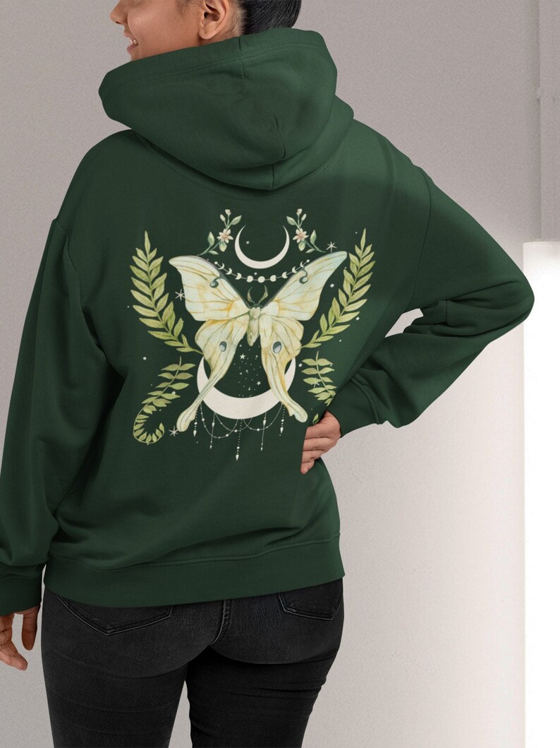 Puede incluir: Una sudadera con capucha verde oscuro con un dise&ntilde;o de polilla luna blanca en la espalda. La polilla est&aacute; rodeada de hojas verdes y una luna creciente con una cadena de cuentas que cuelga.
