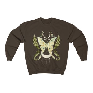 May include: A brown sweatshirt with a green and yellow butterfly design. The butterfly is centered on a crescent moon with a string of beads hanging from it. The design is surrounded by green leaves and flowers.