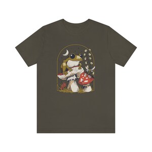 May include: A dark green t-shirt with a graphic of a frog sitting on a mushroom with a smaller mushroom next to it. The graphic is surrounded by a gold frame and has a crescent moon in the background.