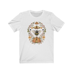 K&ouml;nnte beinhalten: Wei&szlig;es T-Shirt mit einem floralen Kranz-Design mit einer Hummel und zwei Motten. Die Motten sind orange und braun mit schwarzen Markierungen. Die Hummel ist schwarz und gelb.