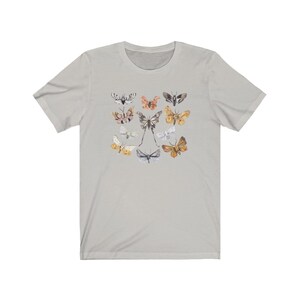 May include: Light gray t-shirt featuring a collection of colorful butterfly illustrations. The butterflies are in various sizes and colors, including shades of brown, orange, white, and gray. The shirt has a crew neck and short sleeves.
