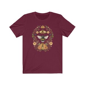 K&ouml;nnte beinhalten: Ein maroonfarbenes T-Shirt mit einem floralen Kranz-Design, das eine Biene und drei Motten zeigt. Der Kranz besteht aus kleinen gelben Bl&uuml;ten, gr&uuml;nen Bl&auml;ttern und wei&szlig;en Punkten.