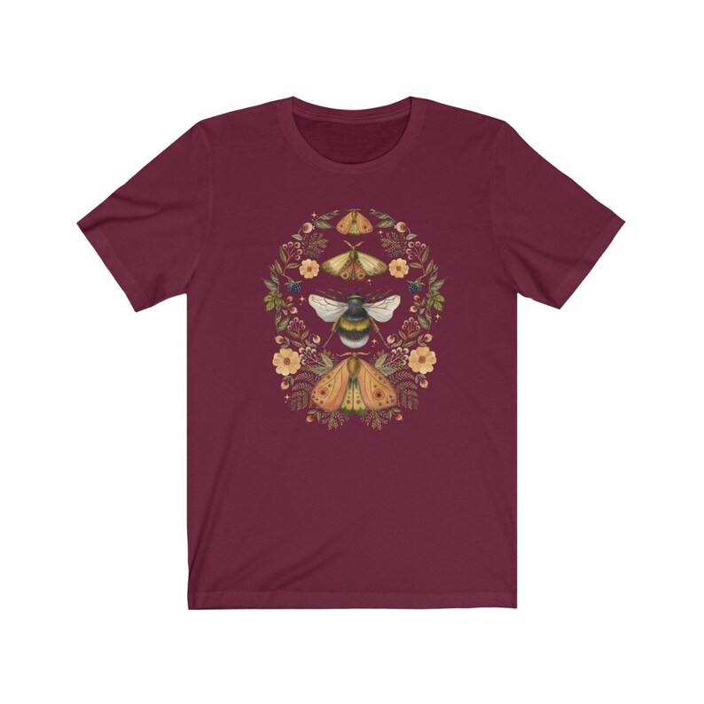 K&ouml;nnte beinhalten: Ein maroonfarbenes T-Shirt mit einem floralen Kranz-Design, das eine Biene und drei Motten zeigt. Der Kranz besteht aus kleinen gelben Bl&uuml;ten, gr&uuml;nen Bl&auml;ttern und wei&szlig;en Punkten.