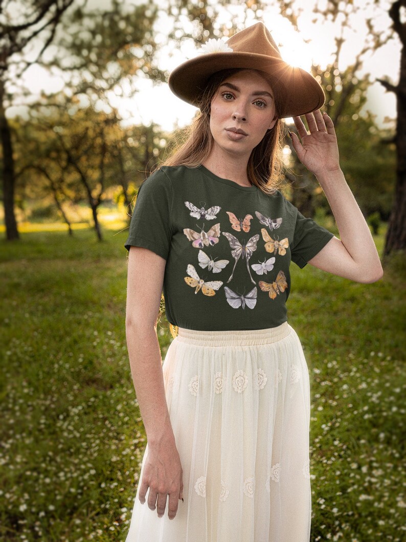 May include: A woman wearing a dark green t-shirt with a butterfly graphic and a cream-colored skirt. She is wearing a brown hat. The background is a green field with trees.