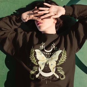 May include: A brown sweatshirt with a green and white moth and moon design. The moth has a crescent moon shape on its wings and is surrounded by green leaves and flowers.