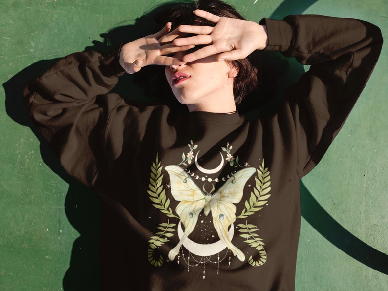 May include: A brown sweatshirt with a green and white moth and moon design. The moth has a crescent moon shape on its wings and is surrounded by green leaves and flowers.