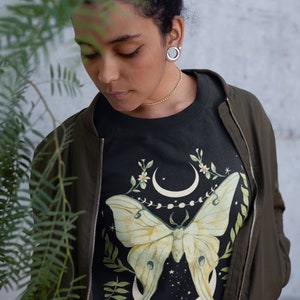 May include: A black sweatshirt with a green and yellow moth illustration. The moth is centered on the shirt and is surrounded by a crescent moon, stars, and floral accents.