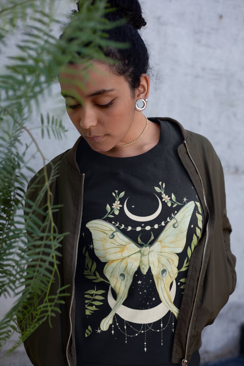 May include: A black sweatshirt with a green and yellow moth illustration. The moth is centered on the shirt and is surrounded by a crescent moon, stars, and floral accents.