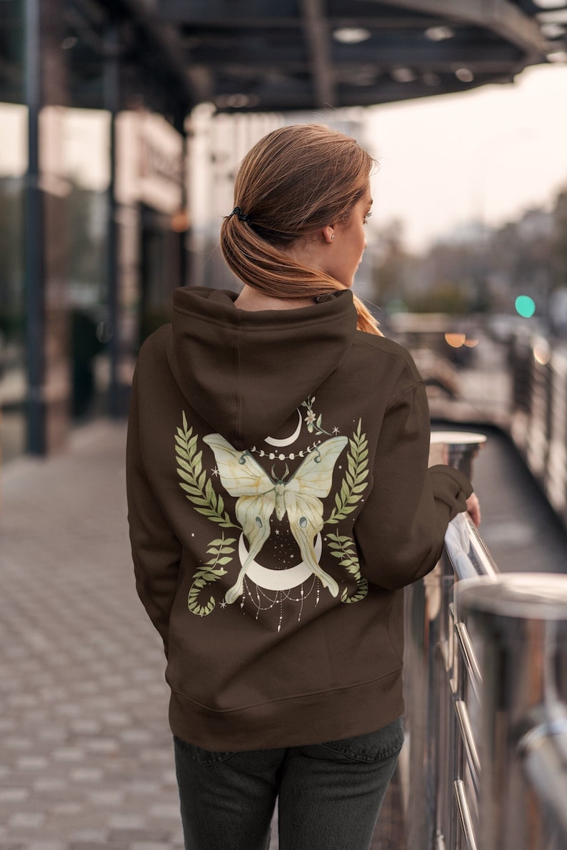 Puede incluir: Una sudadera marr&oacute;n con un estampado gr&aacute;fico de una polilla blanca con hojas verdes y una luna creciente en la espalda.