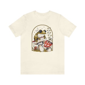 May include: A beige t-shirt with a graphic of a green frog sitting on a mushroom with a red and white mushroom in the background. The graphic is surrounded by a gold frame.