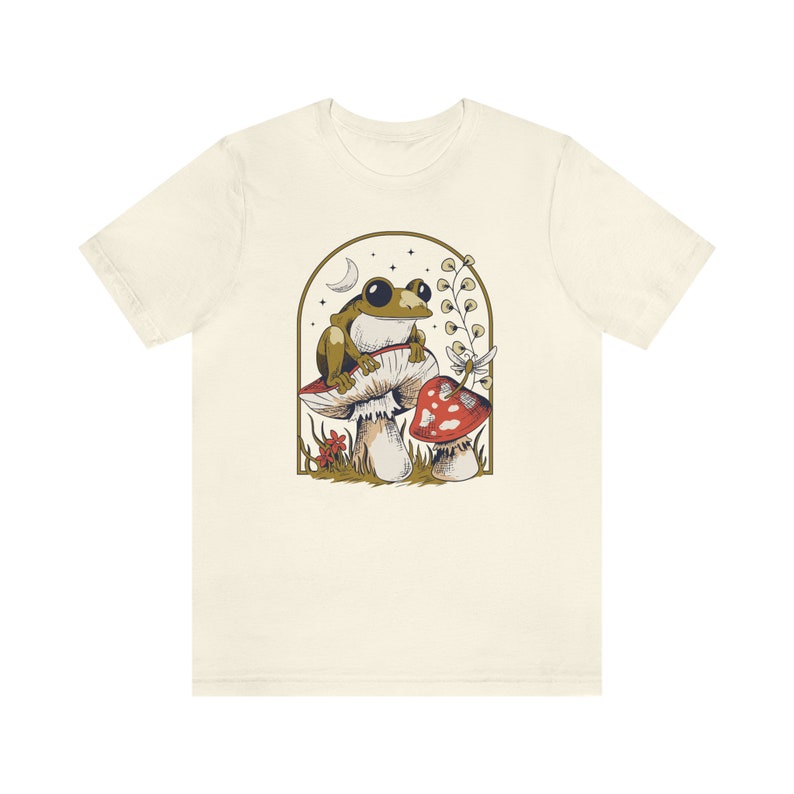 May include: A beige t-shirt with a graphic of a green frog sitting on a mushroom with a red and white mushroom in the background. The graphic is surrounded by a gold frame.