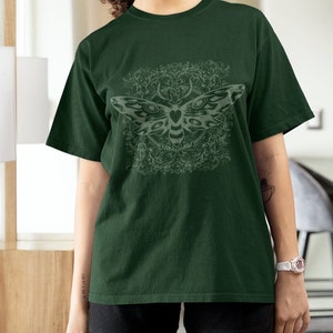 May include: Dark green t-shirt featuring a detailed moth design with a decorative background. The moth is light gray, and the shirt has short sleeves and a relaxed fit. The design is centered on the front of the shirt.