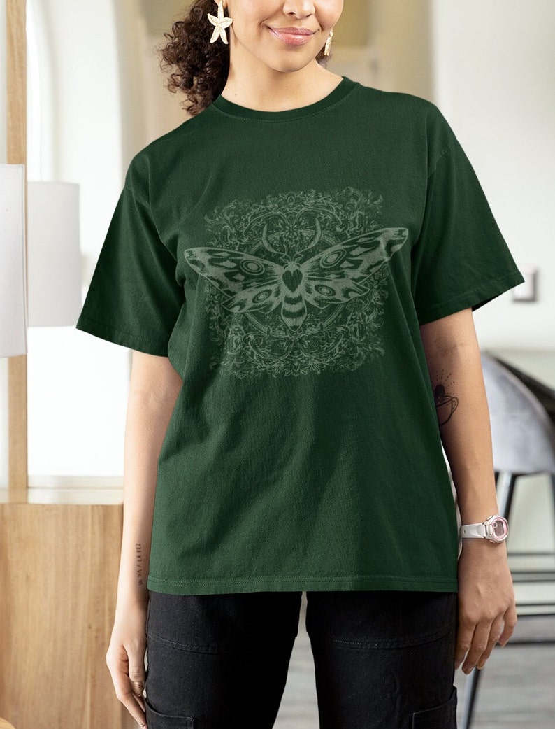 May include: Dark green t-shirt featuring a detailed moth design with a decorative background. The moth is light gray, and the shirt has short sleeves and a relaxed fit. The design is centered on the front of the shirt.