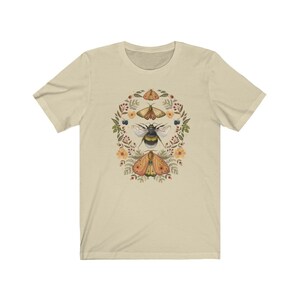 K&ouml;nnte beinhalten: Ein beiges T-Shirt mit einem floralen Kranz-Design, das eine Hummel und drei Motten zeigt. Die Motten sind gelb, orange und braun mit schwarzen Markierungen. Die Hummel ist schwarz und gelb. Die Blumen sind rosa, gelb und wei&szlig;.