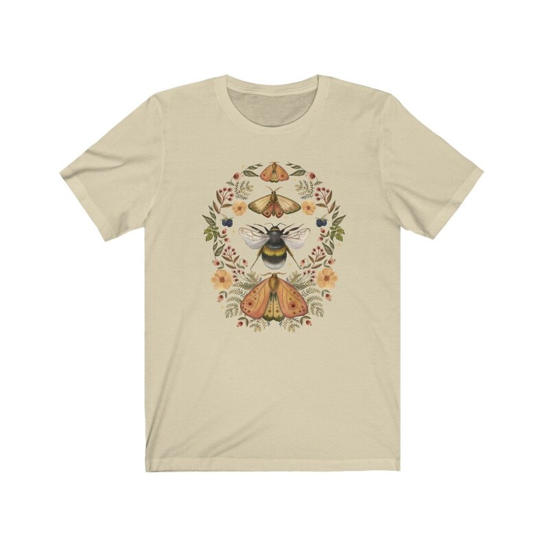 K&ouml;nnte beinhalten: Ein beiges T-Shirt mit einem floralen Kranz-Design, das eine Hummel und drei Motten zeigt. Die Motten sind gelb, orange und braun mit schwarzen Markierungen. Die Hummel ist schwarz und gelb. Die Blumen sind rosa, gelb und wei&szlig;.