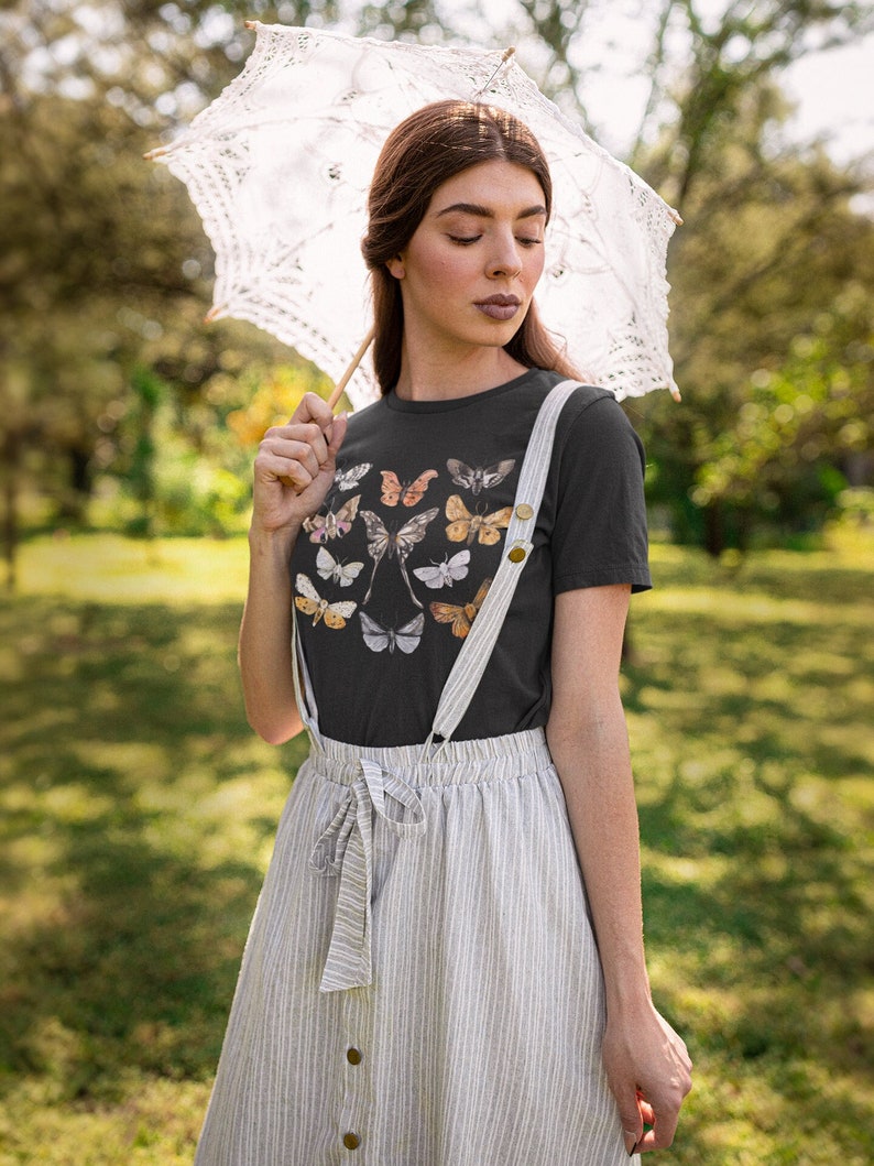 May include: A black t-shirt featuring a collection of butterfly illustrations in various colors. The person is wearing a striped skirt with suspenders and holding a white lace parasol. The background is a blurred green outdoor scene.