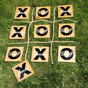 May include: A wooden tic-tac-toe game with the names "Jenna" and "Geoff" written on each square. The game is set up on a green grassy surface.