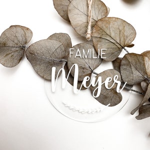 Könnte beinhalten: Ein runder, transparenter Acryl-Anhänger mit dem Text "FAMILIE Meyer" in weißen Buchstaben. Der Anhänger ist mit einem kleinen weißen Blumenmuster verziert und hat eine Schnur zum Aufhängen.
