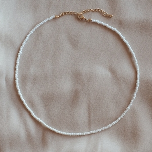 May include: A white beaded necklace with a gold-tone clasp and extender. The necklace is made of small, round white beads. The background is a plain, neutral beige colour.