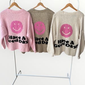 May include: Three sweaters in pink, tan, and cream, each with a large pink smiley face and the words "Have A Good Day!" in black. The sweaters are hanging on wooden hangers against a white background.
