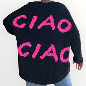 May include: Black knit sweater with the word "CIAO" in bright pink lettering across the back. The sweater has a loose fit and long sleeves. The neckline has a distressed detail.