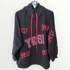 以下が含まれることがあります： ラインストーンの文字で「YES！」、ピンクのスクリプトで「yes！」と書かれた黒いパーカー。 パーカーにはドローストリングが付いており、ハンガーに吊るされています。