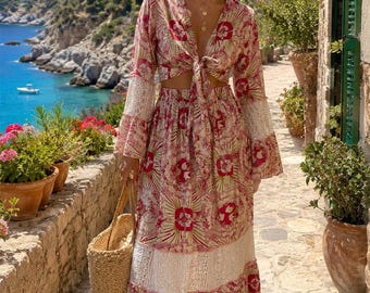 May include: A two-piece set consisting of a long-sleeved tie-front crop top and a matching maxi skirt. The garment features a red and beige floral pattern with white lace trim inserts on the sleeves and skirt, paired with a woven straw tote bag.