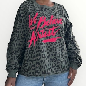 May include: An olive green sweatshirt with a leopard print pattern and the text "We Believe the Artist" in bright pink. The long-sleeved top has a crew neck and ribbed cuffs.