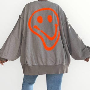 May include: Oversized gray sweatshirt featuring a large, bright orange smiley face design on the back. The sweatshirt has a relaxed fit, long sleeves, and a ribbed hem. The model is wearing blue jeans.