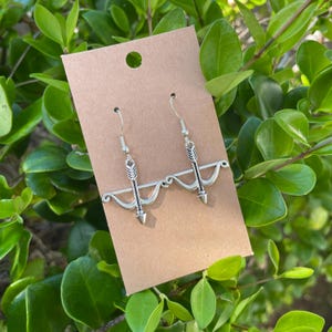 May include: Silver-tone drop earrings with a bow and arrow motif. The earrings are displayed on a brown card, with a background of green foliage. The arrowheads are pointing down, and the bow has a curved design.