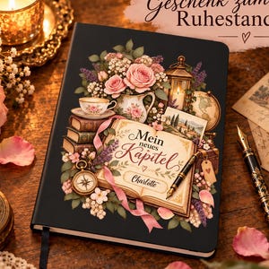 Könnte beinhalten: Ein schwarzes Notizbuch mit Hardcover, verziert mit einer Blumenillustration, Bücherstapeln, einer Laterne, einem Kompass und einem aufgeschlagenen Buch mit dem Text "Mein neues Kapitel" und dem Namen "Charlotte", inklusive Füllfederhalter.
