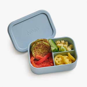 May include: A blue silicone lunch box with three compartments. The box is filled with a variety of snacks, including green pancakes, red peppers, cheese cubes, cucumbers, and yellow crackers. The lid of the box has the name "Noah" written on it.