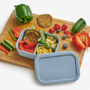 May include: A blue silicone lunch box with a divided interior, filled with snacks like cucumber, red peppers, cheese cubes, and green fritters. The lid has the name "Noah" printed on it.