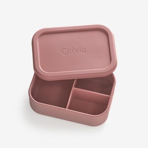 May include: A pink silicone lunch box with three compartments and a lid that has the name "Olivia" embossed on it.