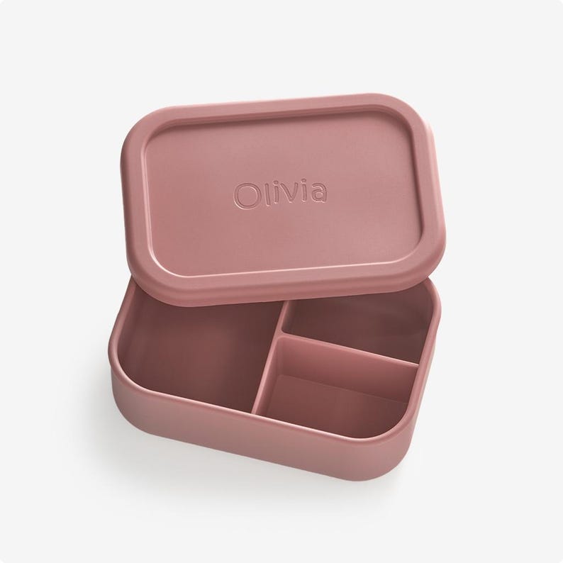May include: A pink silicone lunch box with three compartments and a lid that has the name "Olivia" embossed on it.