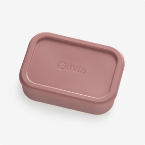 May include: A dusty rose rectangular silicone container with the name "Olivia" embossed on the lid.