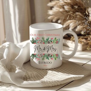 May include: White ceramic mug with the text "Our First Christmas" in red, "Mr & Mrs" in black script, and the last name "Estredo" with the year "2024". The mug is decorated with festive greenery and berries.