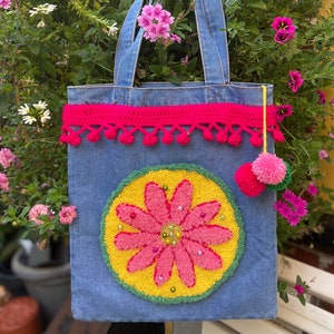 Puede incluir: Una bolsa de mano de mezclilla con un borde de crochet rosa y un diseño de flor rosa en la parte delantera. La flor está rodeada de un círculo amarillo y un borde verde. La bolsa tiene un pompón rosa en el asa.