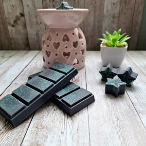 May include: Two black wax melts with green glitter, stacked on a white wooden surface. There are three black star-shaped wax melts on the surface, and a pink ceramic wax warmer with a heart design in the background.