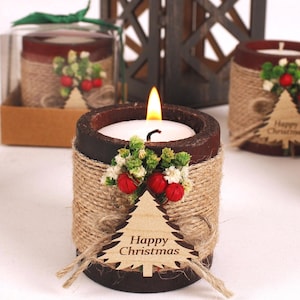 Puede incluir: Un conjunto de portavelas cilíndricos de color marrón oscuro envueltos en cuerda de yute natural, cada uno decorado con una pequeña etiqueta de madera en forma de árbol de Navidad con la inscripción Happy Christmas, bayas rojas y follaje verde.