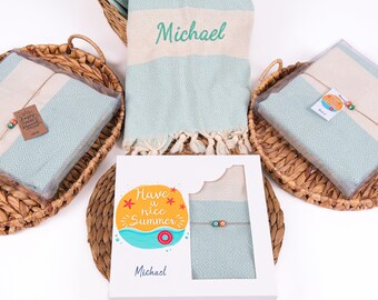May include: A collection of beach towels in a basket, some packaged in clear plastic. The towels are light blue with a diamond pattern and a cream-colored stripe. One towel is embroidered with the name "Michael". The packaging includes a gift box with the text "Have a nice Summer".